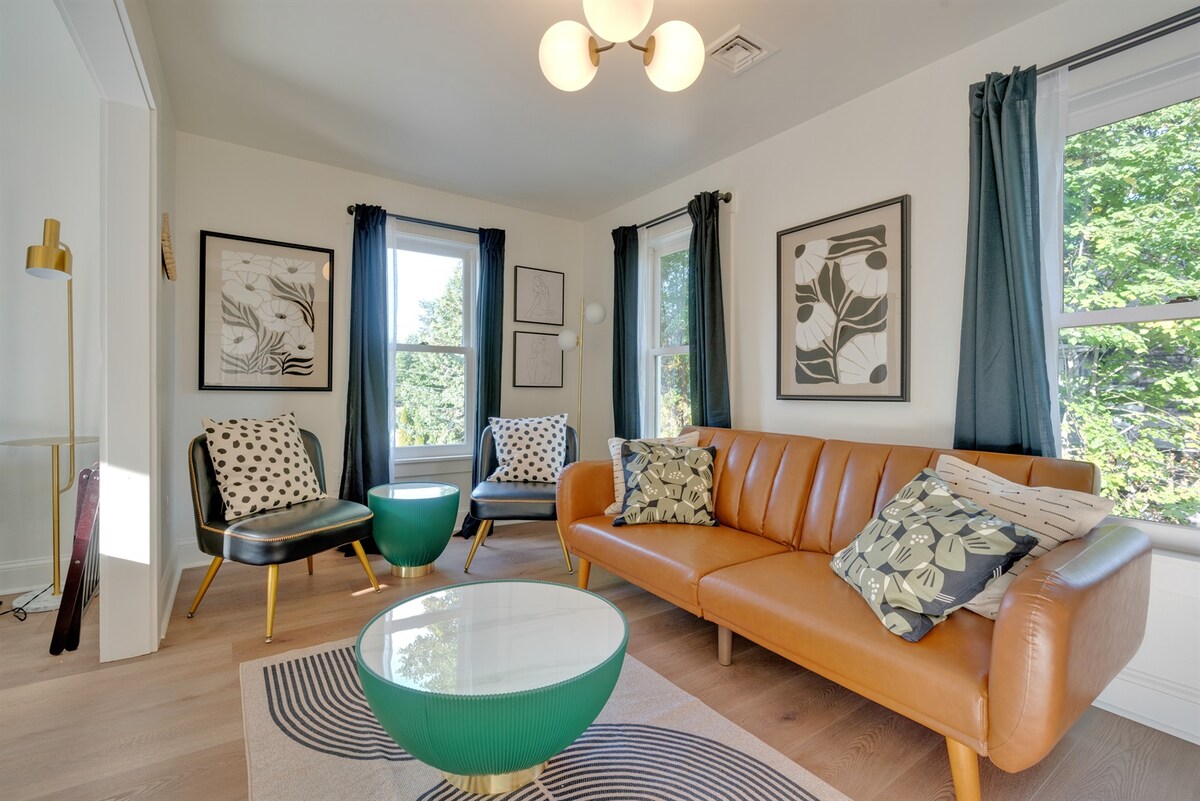 A welcoming living area is illuminated by a modern light fixture. A brown leather sofa is paired with chairs featuring black and white leaf patterns. Two large windows provide natural light, and a round glass coffee table rests on a patterned rug.