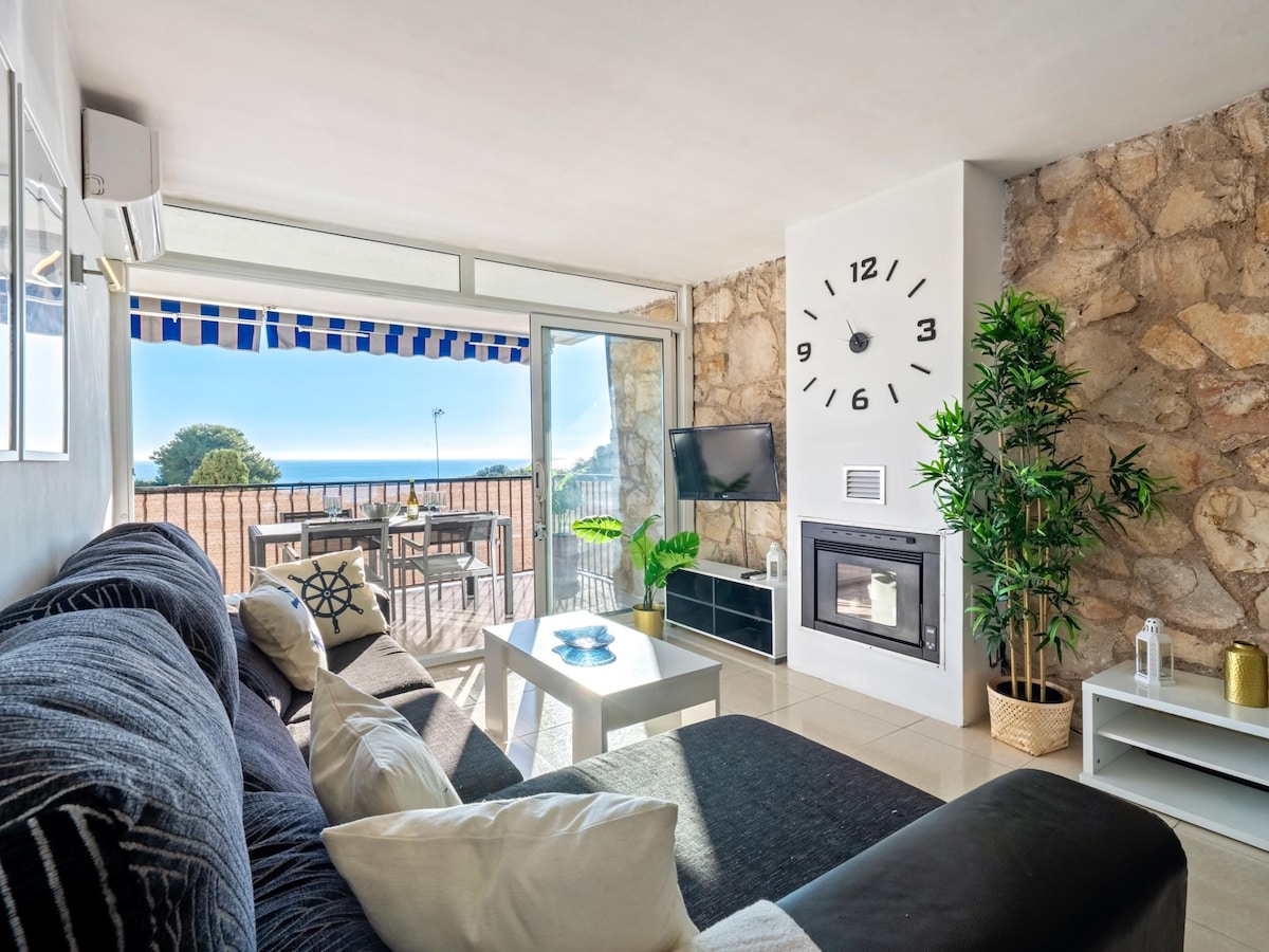 A bright living area features a sectional sofa and a modern coffee table. Large glass doors open to a terrace with outdoor seating, offering views of the sea. A wall clock adds a touch of elegance, and a flat-screen TV is positioned nearby.