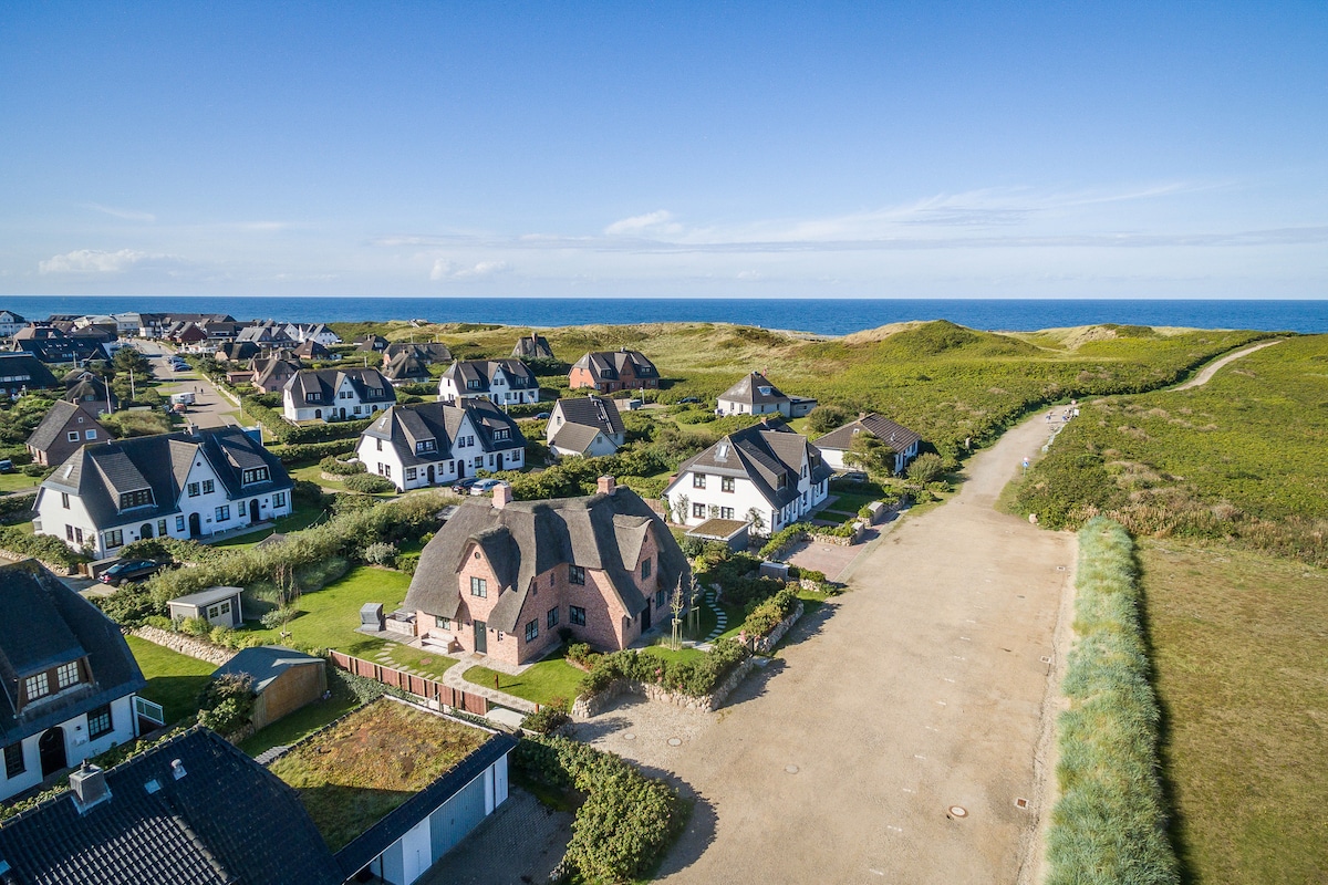 Aerial view of a charming coastal neighborhood featuring house rooftops beneath thatched roofs, with vibrant green landscapes leading to dunes in the background. The ocean is visible on the horizon, highlighting the proximity to the beach.