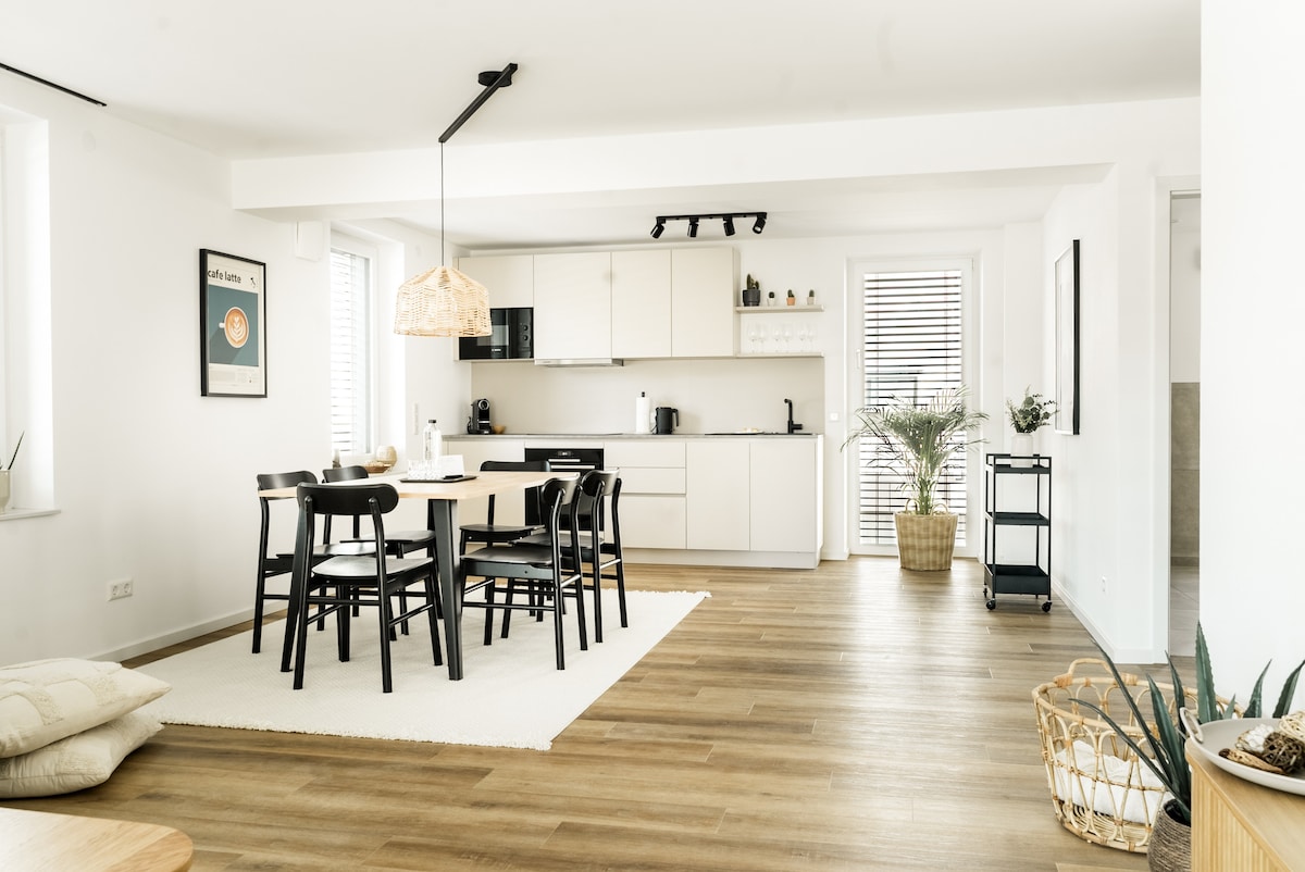 An open-plan living area is featured, showcasing a dining table surrounded by black chairs. A fully equipped kitchen is visible, with modern appliances and cabinetry. Light wood flooring complements the neutral color palette, while natural light streams in through multiple windows.
