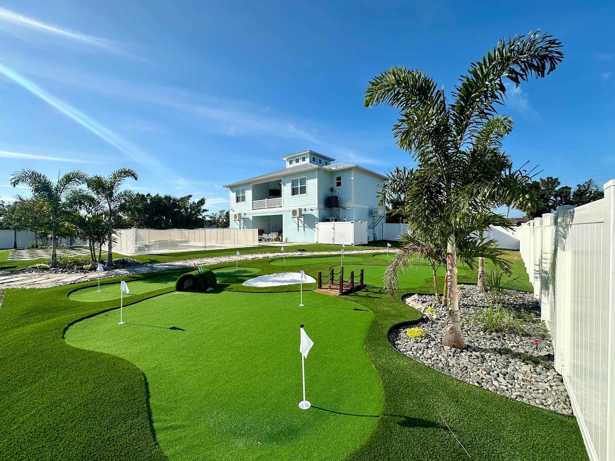 A well-maintained outdoor space features a private mini golf course with three putting greens and surrounding palm trees. The Key West-style home is visible in the background against a clear blue sky, with neatly trimmed landscaping and a fenced yard enhancing the tranquil setting.