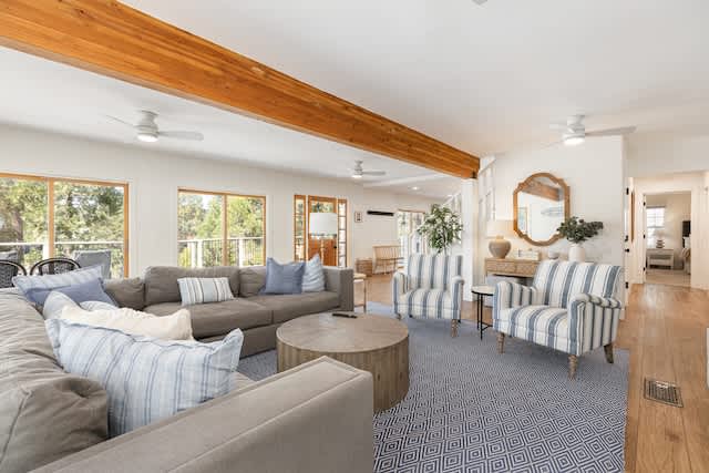A spacious living area features a large sectional sofa accompanied by two striped chairs. Natural light filters through multiple windows, highlighting the wooden beam ceiling. A round coffee table sits centrally on a textured area rug, creating a welcoming gathering space.
