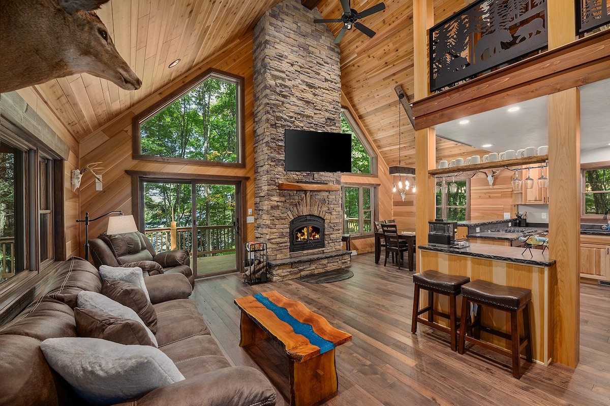 A spacious living area is highlighted by a large stone fireplace and wooden beams above. Ample seating is provided on a plush sofa, and a coffee table with a blue accent runs centrally. Large windows frame the views of the surrounding trees, allowing natural light to pour in.
