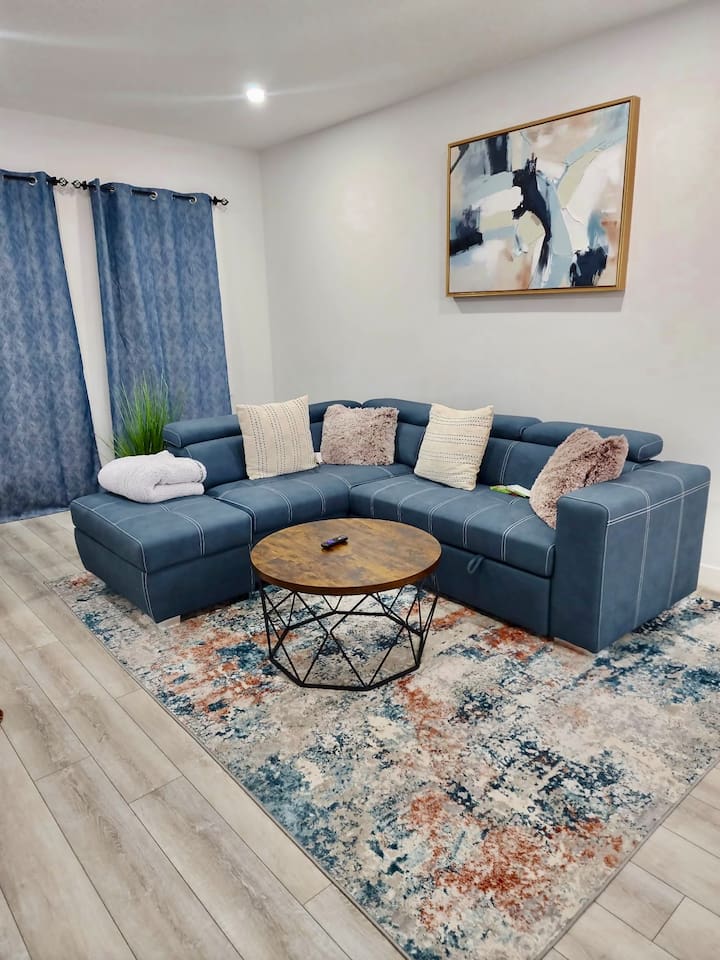 A comfortable living area features a sectional sofa adorned with decorative pillows. A round coffee table sits at the center on a patterned area rug, while blue curtains frame the window. Wall art adds a modern touch to the neutral-toned walls.