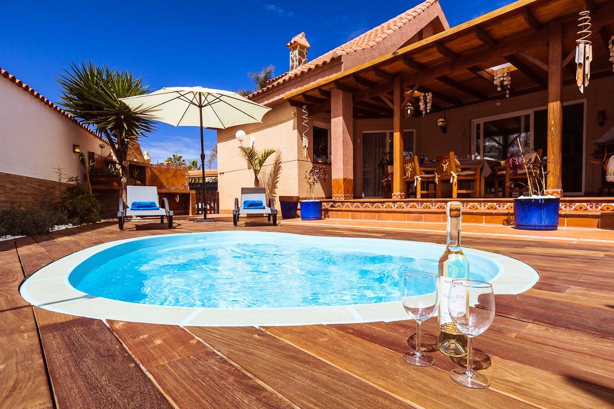 A private shallow pool is surrounded by wooden decking, with two sun loungers resting nearby. An umbrella offers shade, and a bottle of wine paired with two glasses is positioned on the deck, creating a welcoming outdoor space for relaxation and enjoyment.