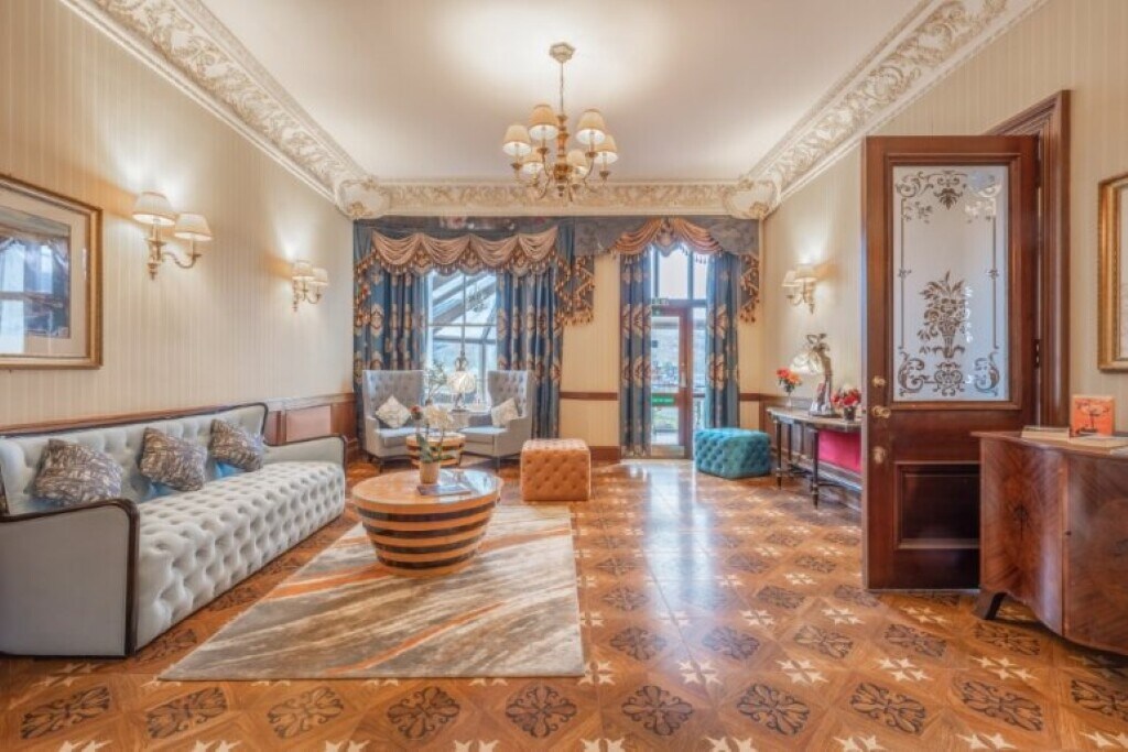 A well-appointed lounge area features elegant lighting and intricate ceiling details. Plush sofas and an ornate coffee table are arranged on a decorative floor. Large windows allow natural light to illuminate the space, which is enhanced by rich drapes and tasteful decor.