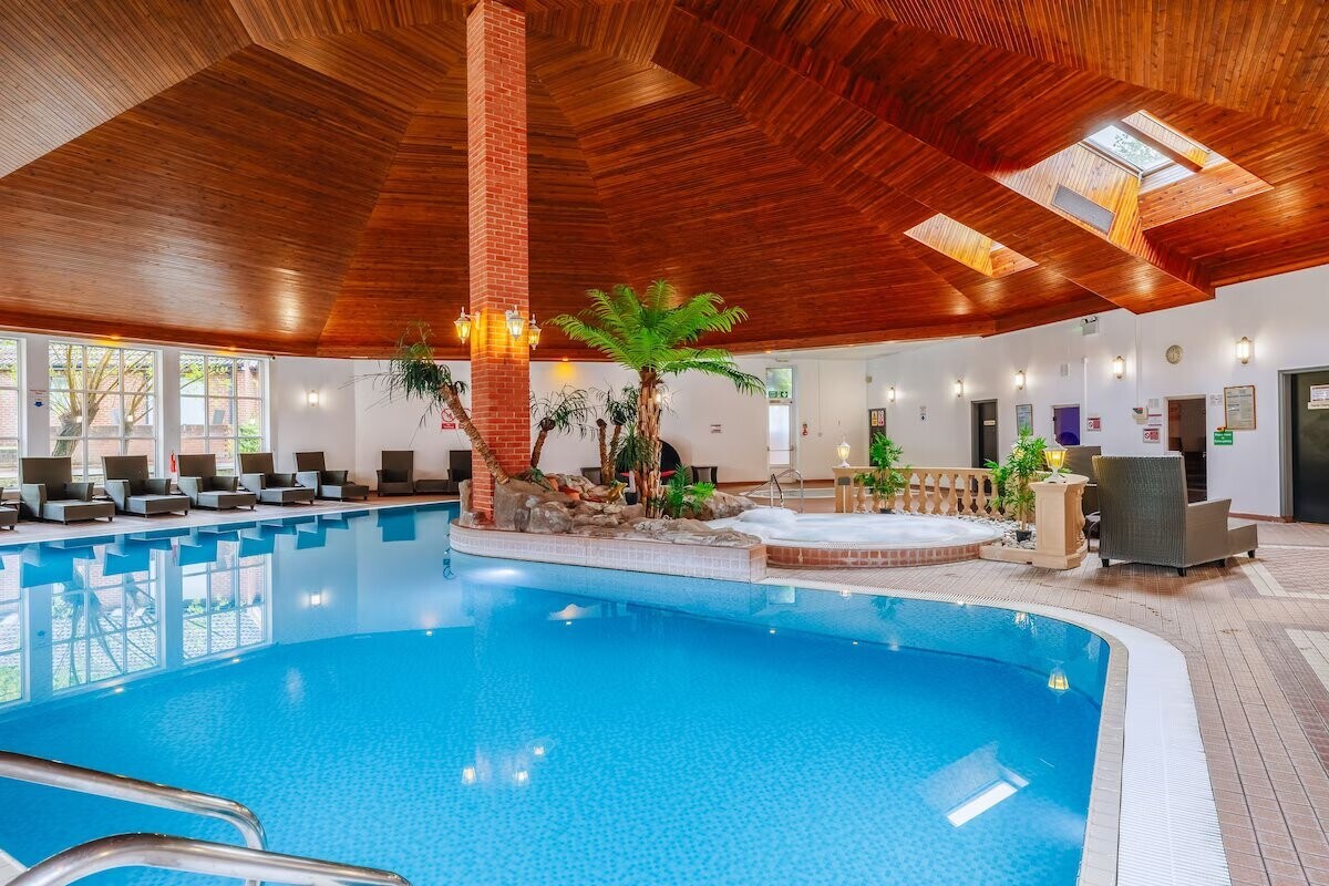 An indoor swimming pool is surrounded by elegant loungers, featuring a soothing atmosphere created by natural light filtering through skylights. The pool area showcases a blend of wood and stone accents, complemented by lush greenery. A nearby hot tub offers a relaxing retreat.