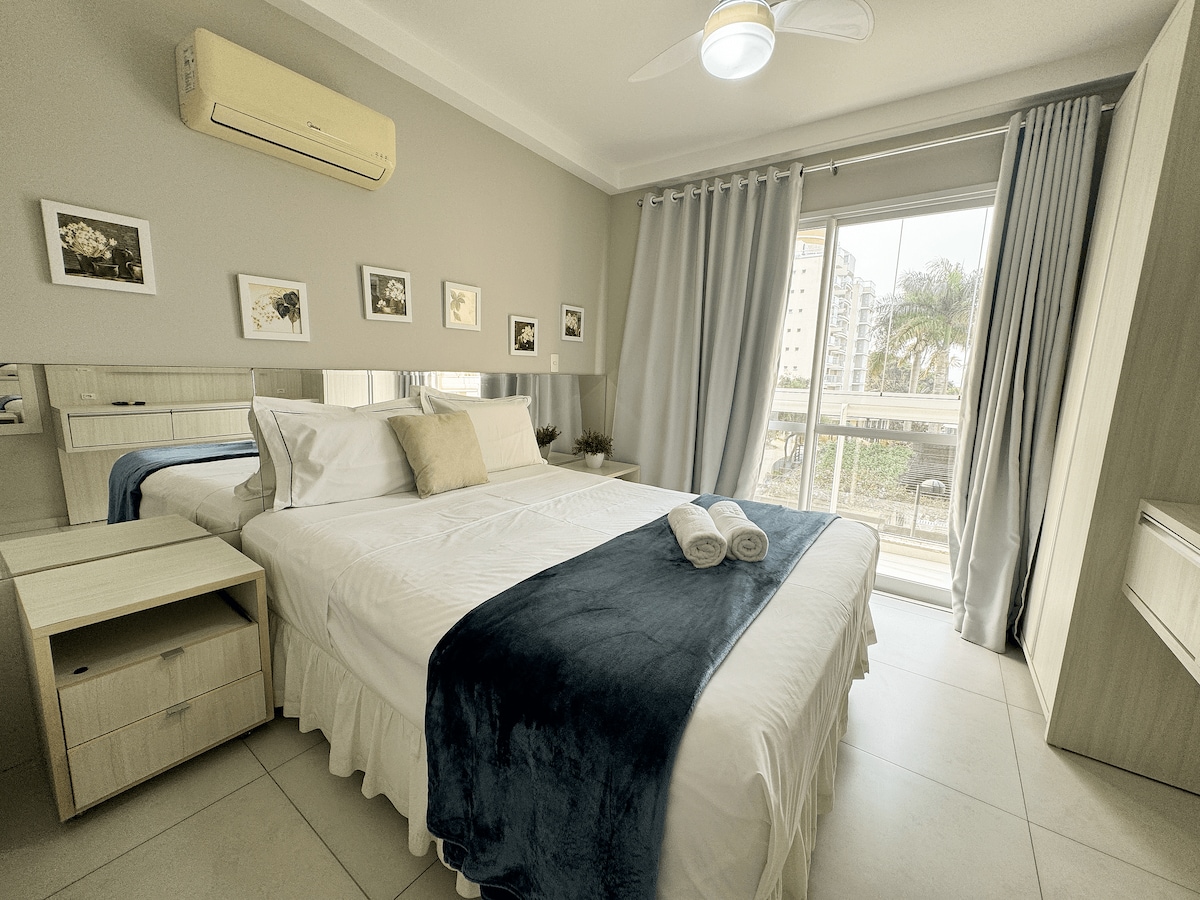 A master suite is depicted with a spacious queen bed, flanked by bedside tables. Natural light filters through a balcony door, complemented by light gray curtains. A ceiling fan and air conditioning enhance comfort. Wall art adds visual interest to the neutral decor.