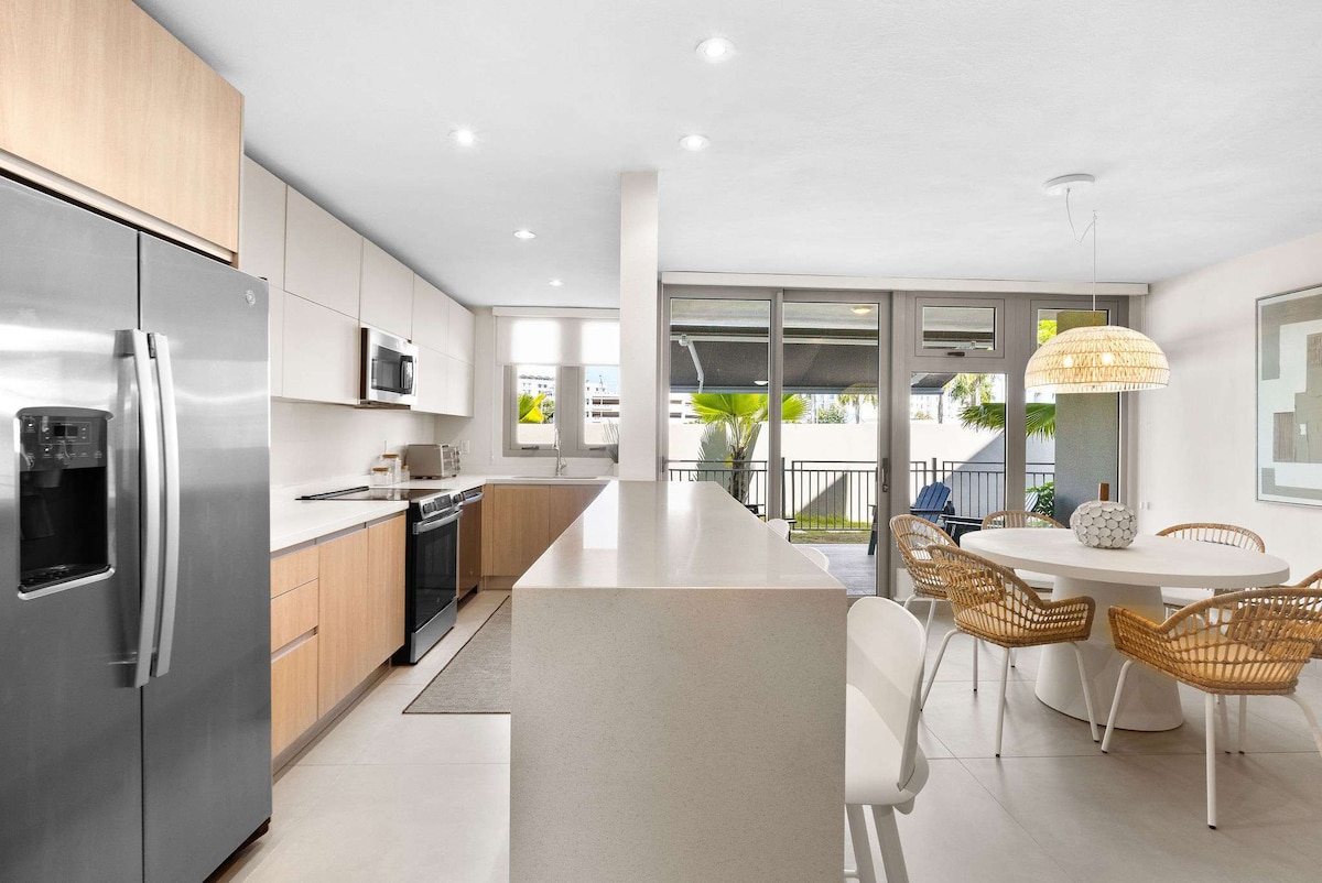 The modern kitchen features sleek cabinetry and stainless steel appliances, including a double-door refrigerator and built-in microwave. An island with seating for four transitions into a dining area with a round table surrounded by rattan chairs. Large windows invite natural light and offer views of the outdoor space.