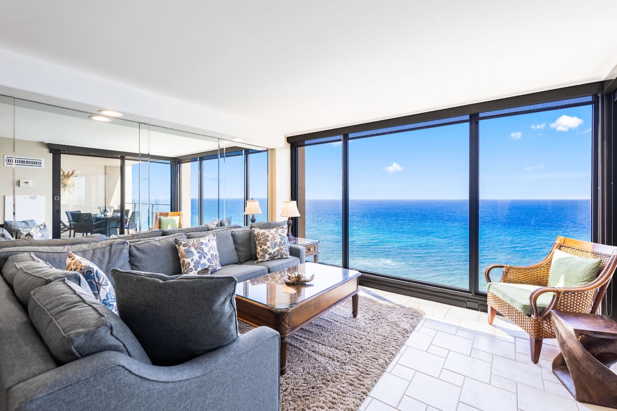 The living area features large windows offering unobstructed ocean views. A spacious gray sectional sofa surrounds a wooden coffee table, complemented by a woven chair. The room is illuminated by natural light, enhancing the light-colored tile flooring and reflecting off the mirror-backed wall.