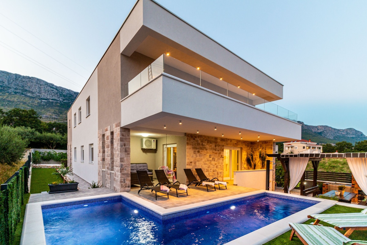 A contemporary two-story house is captured during twilight, featuring a serene pool surrounded by lounge chairs. A balcony extends over the entrance, providing additional space for relaxation. The natural landscape enhances the setting with mountains visible in the background.
