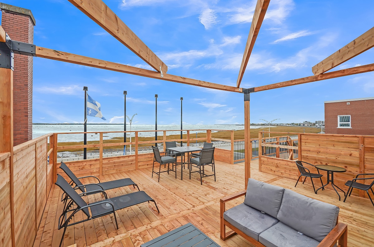 The rooftop terrace is a spacious wood deck featuring multiple seating areas, including lounge chairs and a dining table. An expansive view of the surrounding area and river is visible, with flags gently waving in the breeze. The space offers a relaxing outdoor environment.