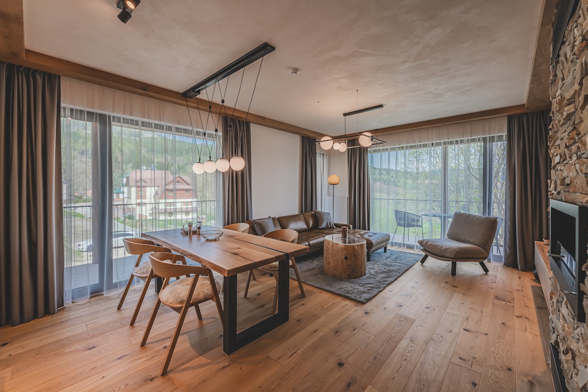 A spacious living room features a wooden dining table accompanied by light-colored chairs. A large, comfortable sofa is arranged around a central coffee table made from natural wood. Floor-to-ceiling windows allow natural light to fill the space, with scenic views visible through sheer curtains.