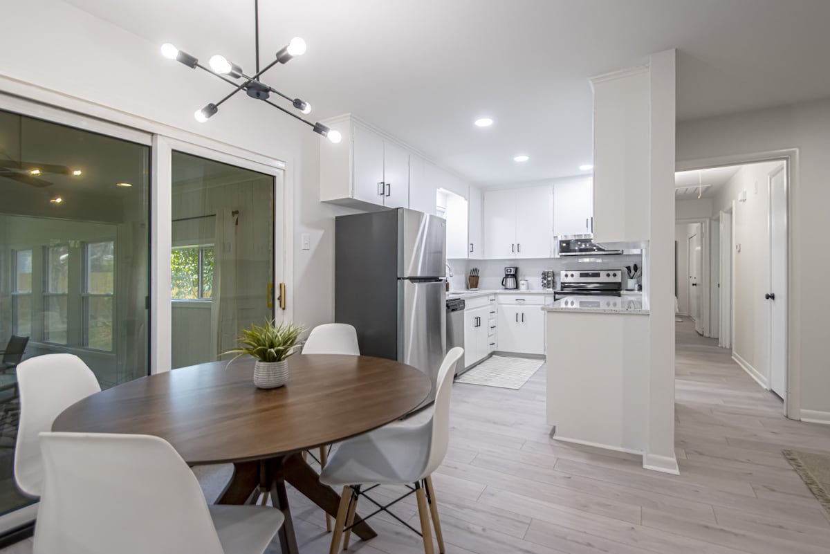 A modern dining area features a round wooden table surrounded by white chairs. Natural light floods in from sliding glass doors leading to a backyard. The kitchen showcases stainless steel appliances and a clean, bright design, contributing to an inviting cooking space.