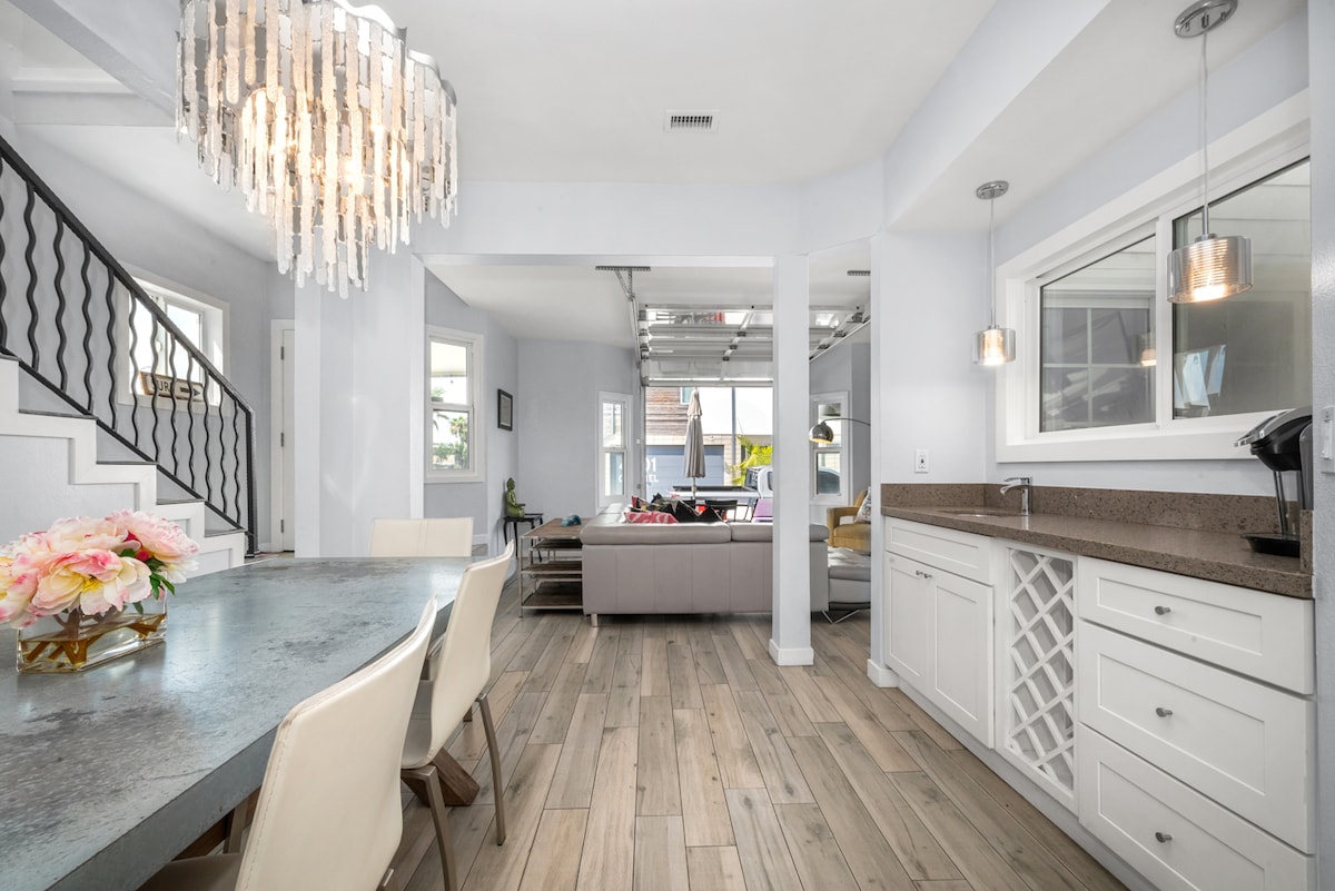 A large dining table is set beneath a modern chandelier, surrounded by light-colored chairs. The open layout connects the dining area to a spacious living area featuring a comfortable couch, while large windows provide ample natural light throughout.