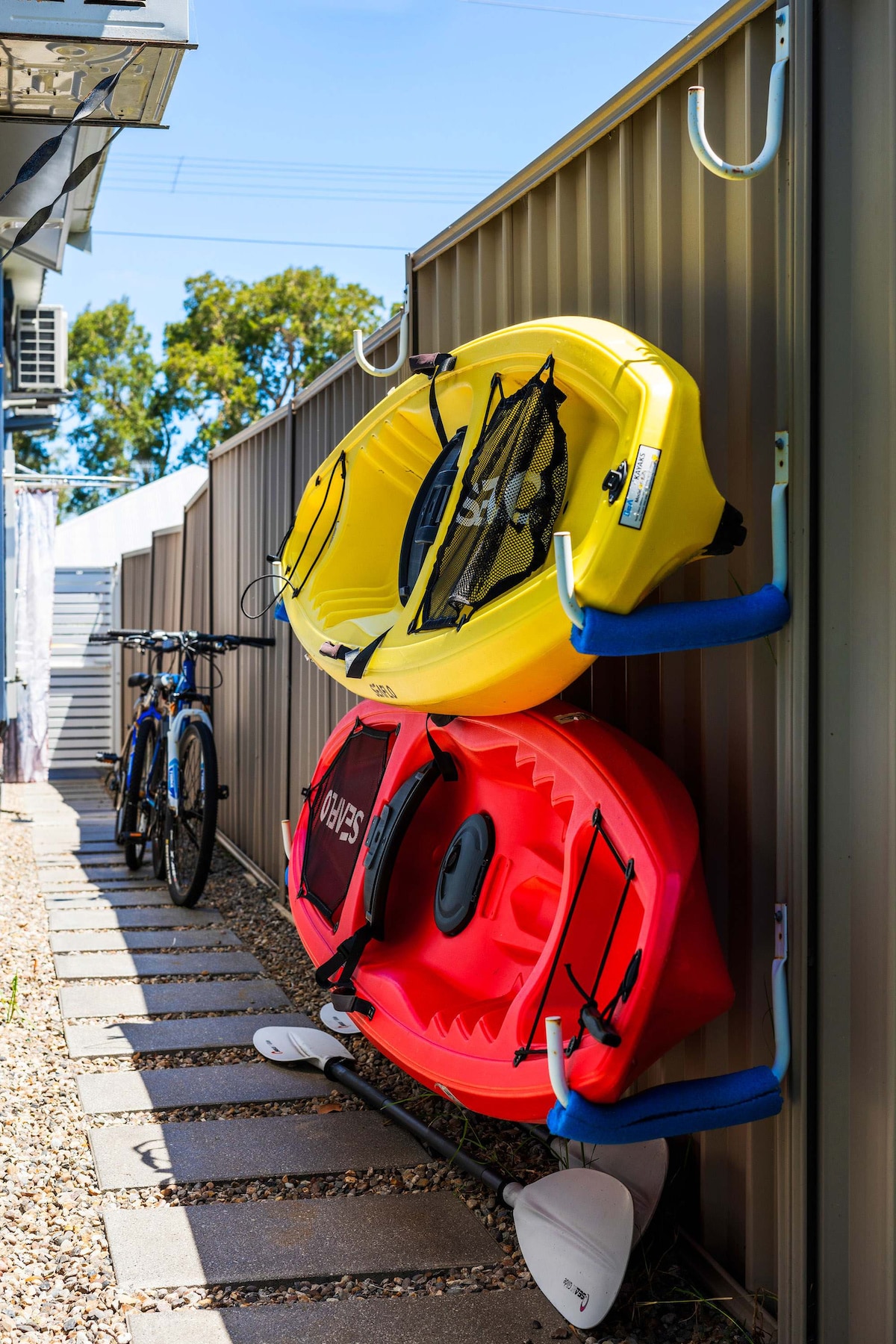 A yellow kayak and a red kayak are securely mounted on a wall, with paddles resting beneath them. Two bicycles are parked alongside the kayaks on a path lined with gravel, indicating a space designed for outdoor activities.