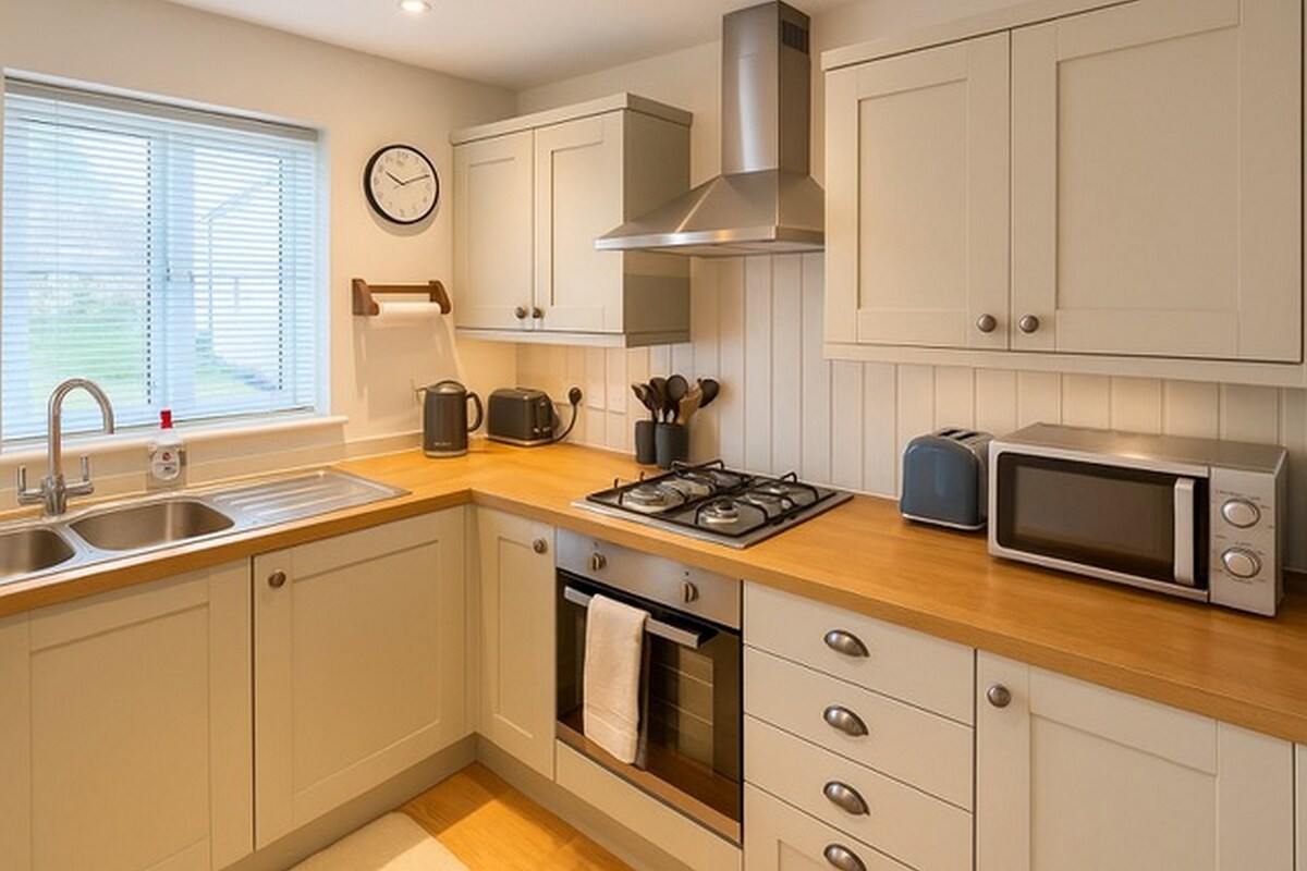 A modern kitchen features a gas hob, electric oven, and microwave within stylish cabinetry. A sink is positioned under a window, providing natural light. Counter space is complemented by wooden surfaces, and kitchen essentials such as a kettle and utensils are neatly arranged for convenience.