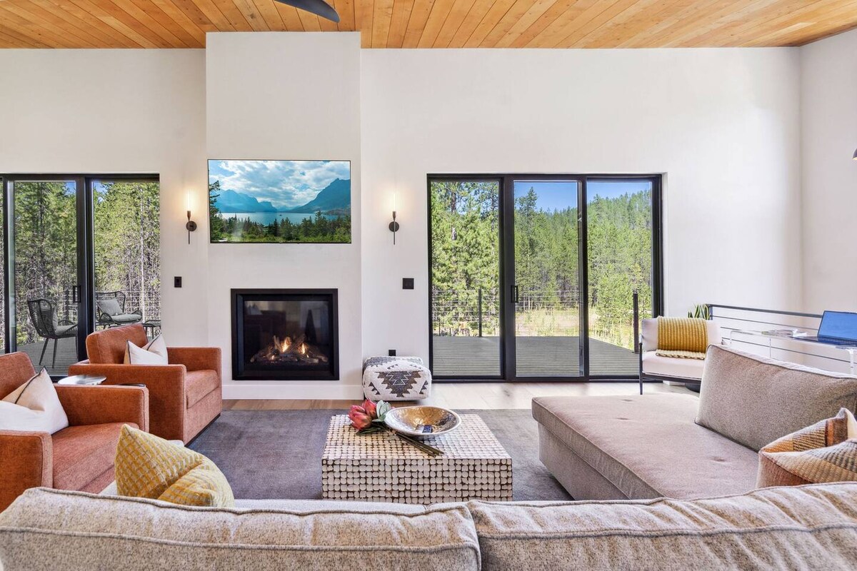 A spacious living room featuring a cozy gas fireplace and large windows that provide views of the surrounding forest. Comfortable seating is arranged around a coffee table, with warm tones and textures highlighted by natural light from outside.