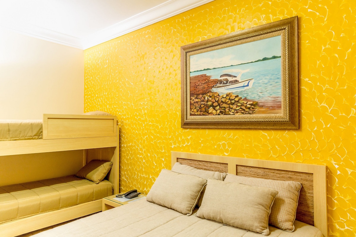 A bright yellow wall features a large painting of a boat on the water, enhancing the room's lively atmosphere. A spacious bed with neutral bedding is positioned alongside a bunk bed, providing ample sleeping arrangement. A bedside table is visible next to the main bed.