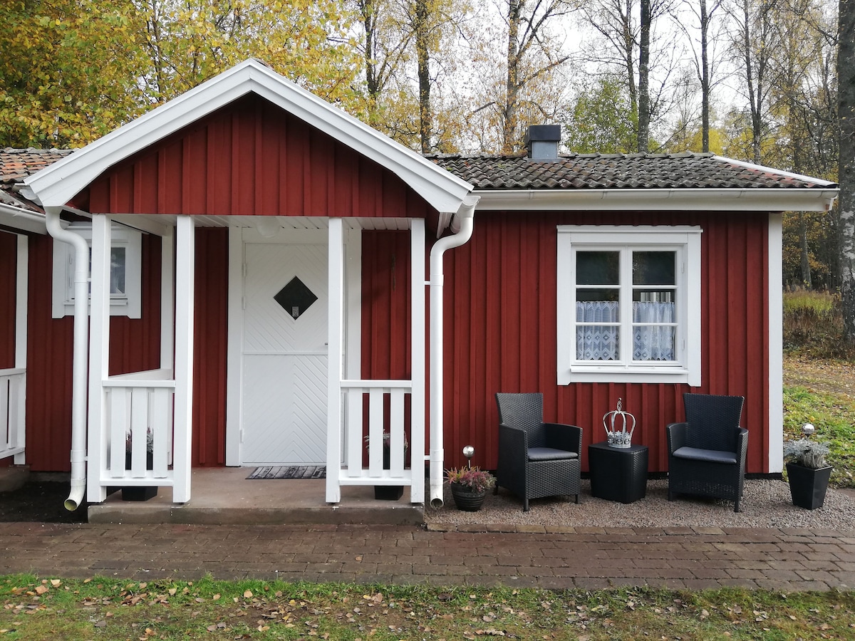A charming red cottage is seen with a white-trimmed porch. Two dark chairs are positioned on the porch, complemented by a potted plant. The entrance features a diamond-shaped window, enhancing the inviting appearance of the home.