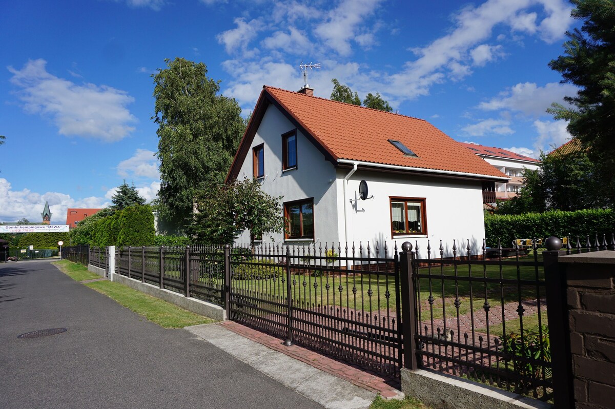 A two-story house is depicted with a red-tiled roof and white exterior walls. Large windows frame the front, surrounded by a well-kept garden and a decorative fence. A gravel pathway leads toward the entrance, set against a clear blue sky with scattered clouds.