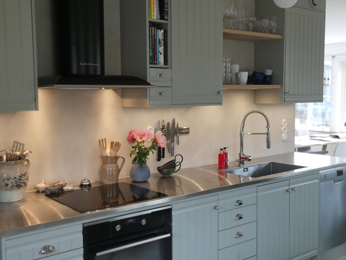The kitchen features light grey cabinetry and stainless steel countertops, creating a functional cooking environment. A bouquet of pink roses sits next to kitchen utensils on the counter, while a modern faucet and cookware complement the space. Open shelves display dishes and glassware.
