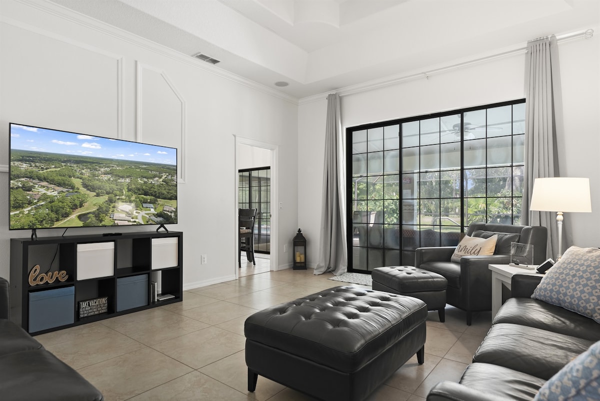A spacious living room features large windows that allow natural light to fill the area. Comfortable black leather seating is arranged around a central ottoman and a television, providing a relaxing environment. A decorative piece on the shelf adds a subtle touch of personalization.
