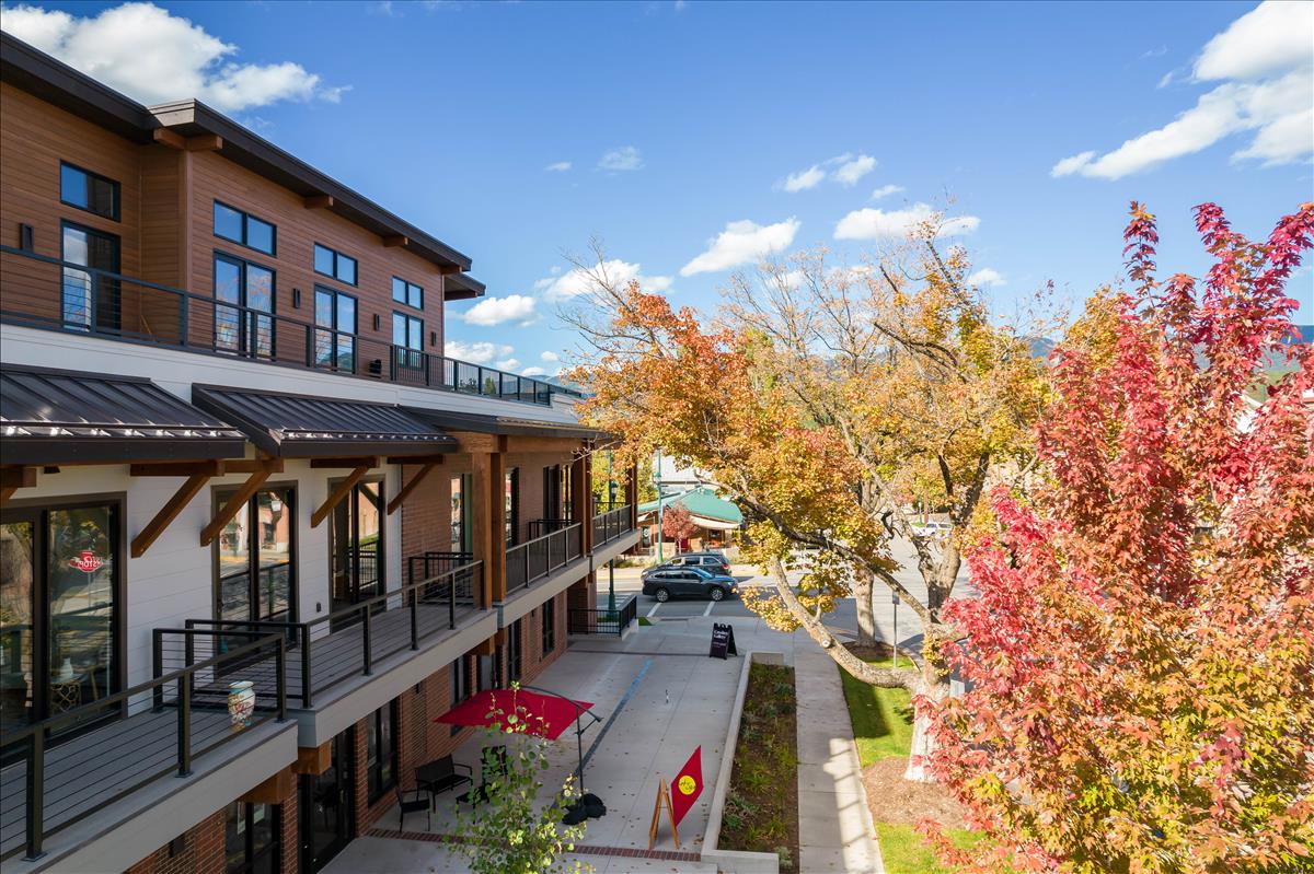 The image showcases a modern building featuring large windows and a welcoming entrance. Vibrant autumn foliage decorates the surrounding area, with a pop of red from the patio umbrellas. Blue skies enhance the inviting atmosphere of this downtown location.