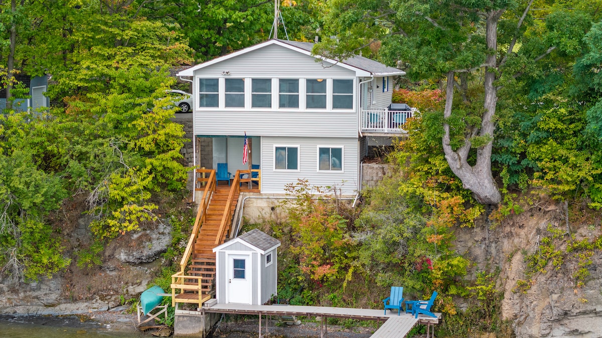 The cottage is nestled among greenery, showcasing a wooden staircase leading from the lakeside dock to the main entrance. Large windows frame the front of the home, offering views of the surrounding nature and water. A small storage shed and seating area are visible near the dock.
