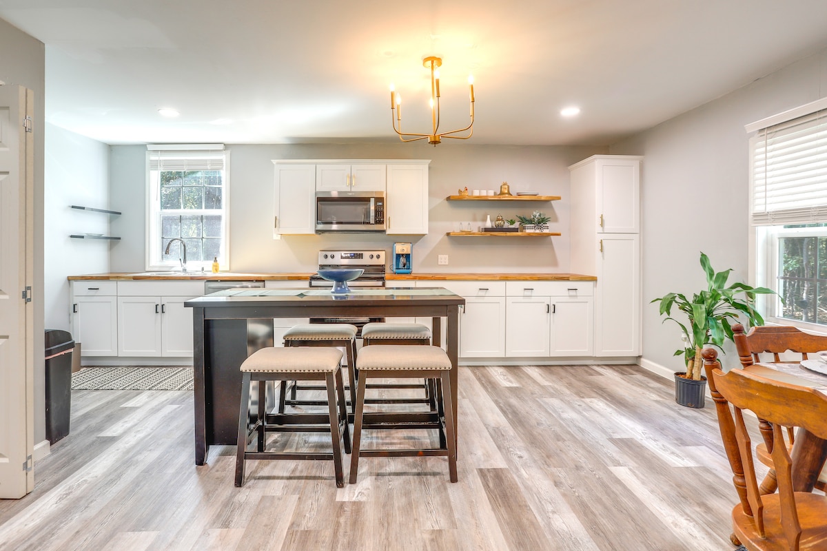 A bright kitchen area features white cabinetry and modern stainless steel appliances. A central island with seating is complemented by a nearby dining table. Natural light flows in through the window, highlighting the sleek flooring and decorative plants in the corner.