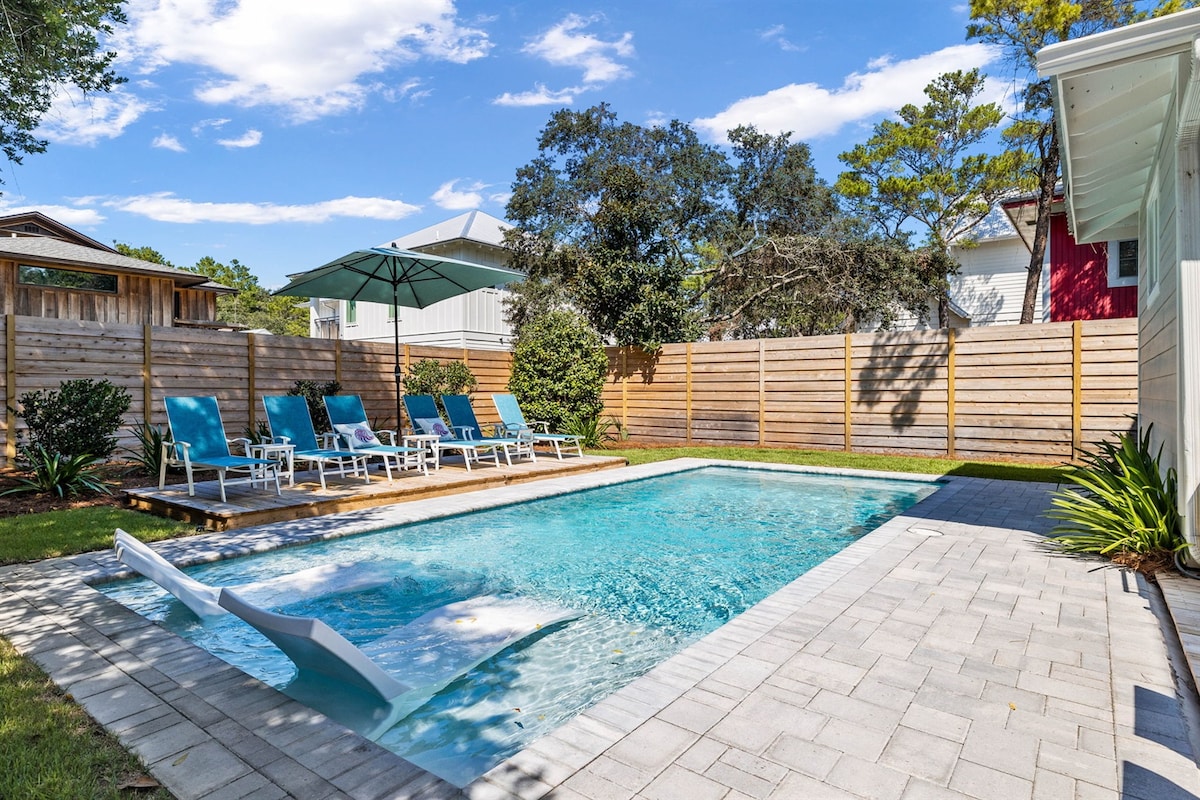 A private pool area is surrounded by a wooden privacy fence. Light blue lounge chairs are arranged on the patio, complemented by a shaded umbrella. The pool's clear water glistens under the sun, with a small wooden deck leading into the pool area.