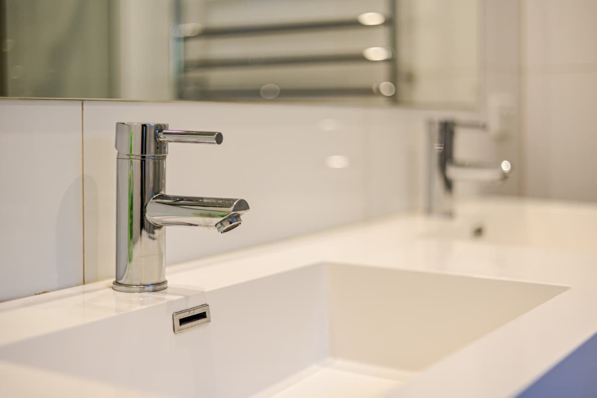 A modern bathroom sink is shown with a sleek chrome faucet positioned above it. The counter features a minimalist design with clean lines, complemented by a large mirror behind. Additional faucets are visible, enhancing the contemporary aesthetic.