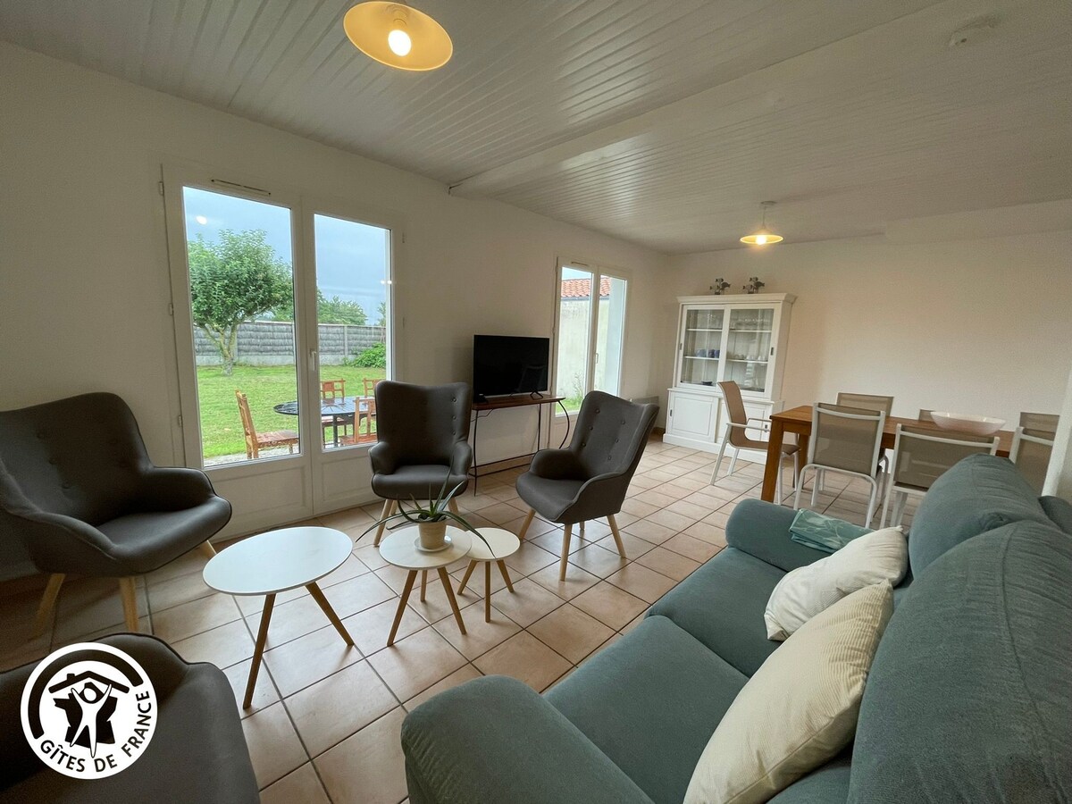 A spacious living area is presented, featuring two sets of comfortable armchairs facing a coffee table. A sofa with soft cushions is positioned against a wall. Large glass doors allow natural light in and provide a view of the garden, where outdoor seating is arranged.