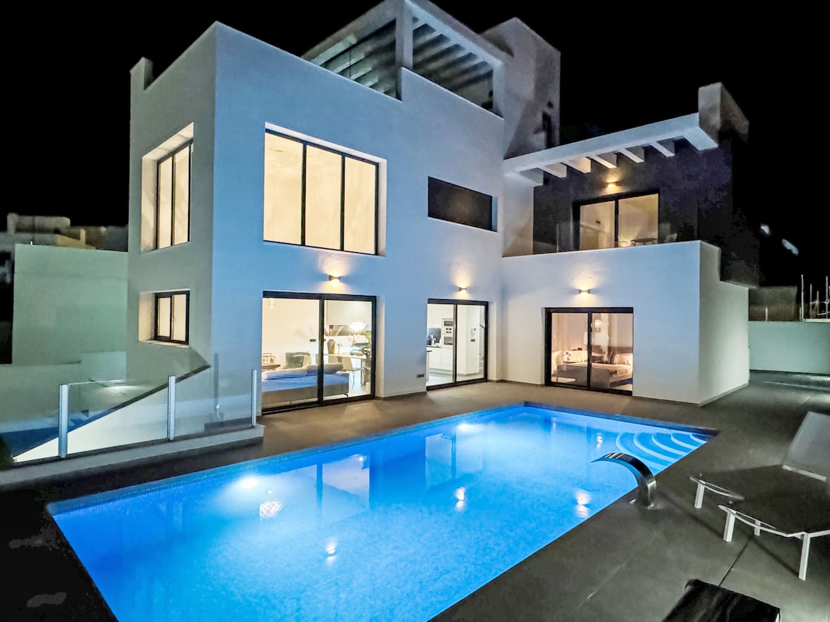 The villa is showcased at night, illuminated by soft exterior lights. The pool reflects a calming blue hue, while large windows reveal a glimpse of the modern interior. Outdoor seating is visible, providing a space to enjoy the serene environment.