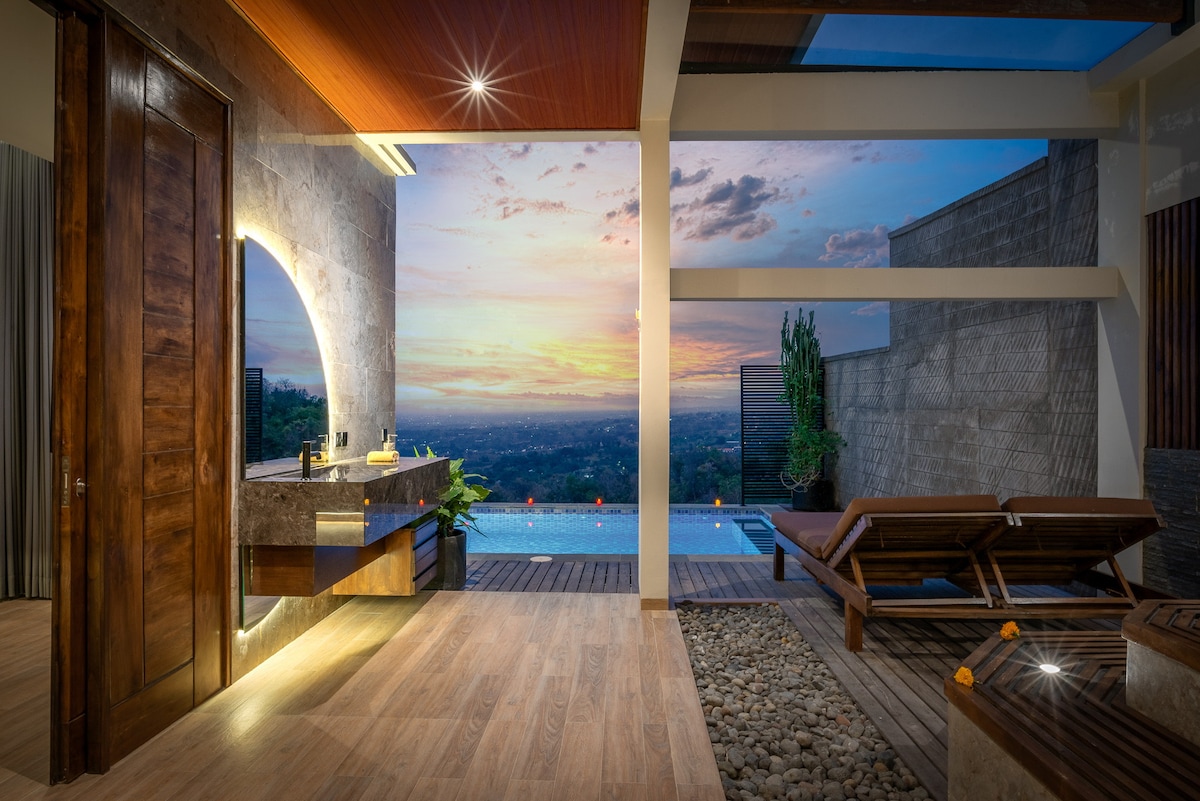 An elegantly designed bathroom features a modern vanity with a large wooden mirror and a view through an arched window. Beyond, the serene outdoor area includes a sunbed and infinity pool, with a vibrant sunset illuminating the horizon.