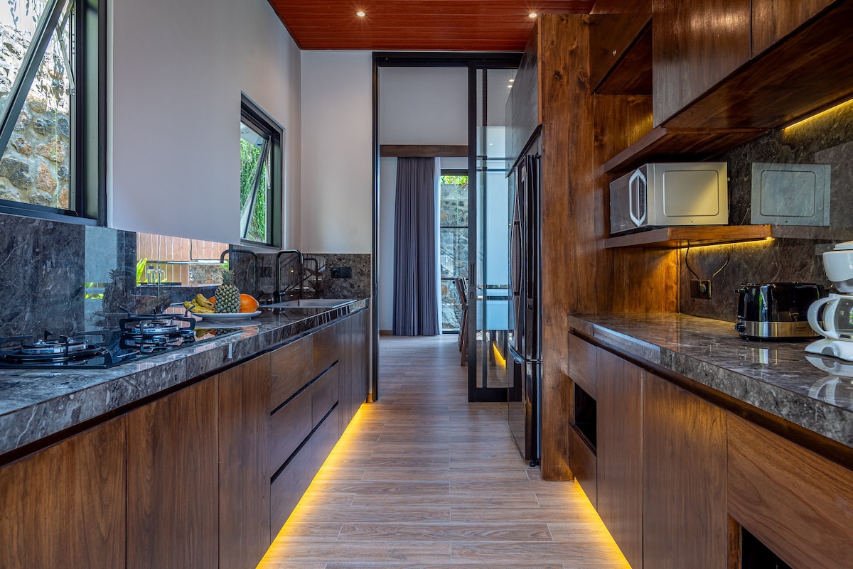 A modern kitchen features sleek cabinetry and dark marble countertops illuminated by warm, under-cabinet lighting. Premium appliances, including an oven and microwave, are visible. Large windows allow natural light to fill the space, providing views of the outdoor area beyond.