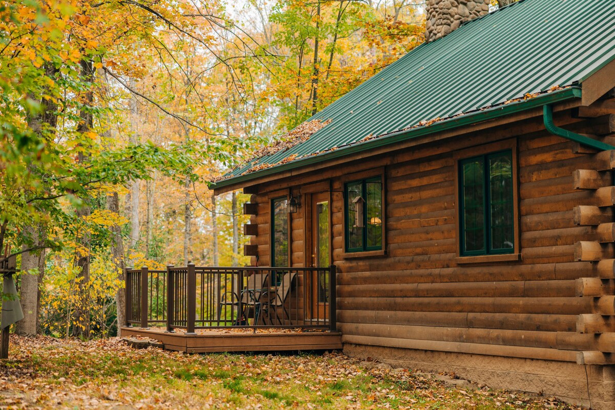 The cabin's exterior showcases natural wood siding complemented by a green metal roof. A private deck with seating is framed by vibrant fall foliage, enhancing the serene atmosphere of the surrounding landscape.