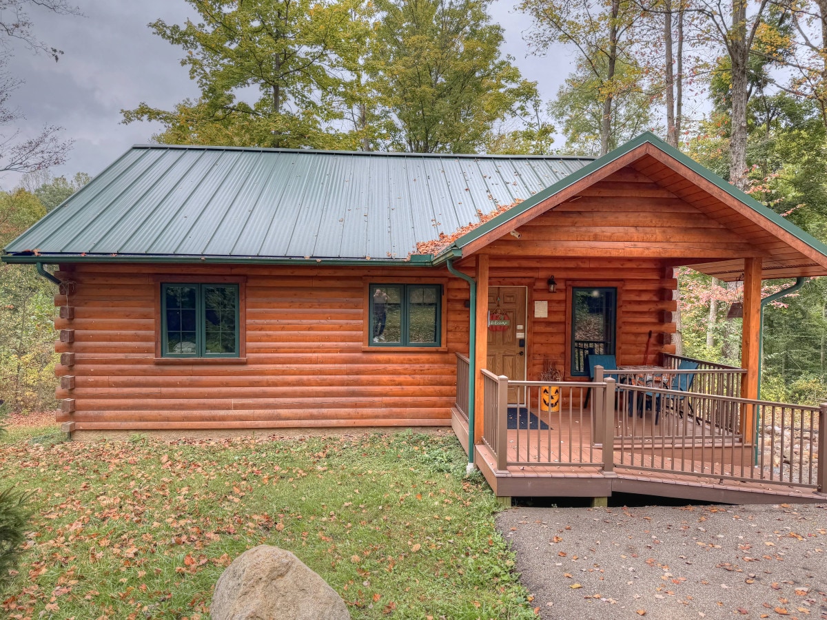 The charming cabin exterior features a rustic log construction, accented by a metal roof. A welcoming front porch is furnished with chairs, providing a comfortable spot to enjoy the surrounding natural landscape, which includes lush trees and a manicured lawn.