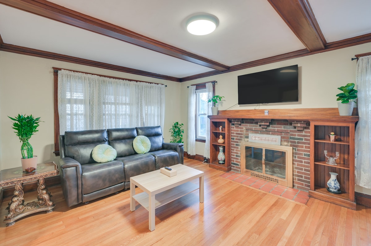 A spacious living area is furnished with reclining seating and a coffee table, complemented by a decorative fireplace. Light-colored curtains frame the windows, and potted plants are strategically placed, adding a touch of greenery. A flat-screen TV is mounted on the wall for entertainment.