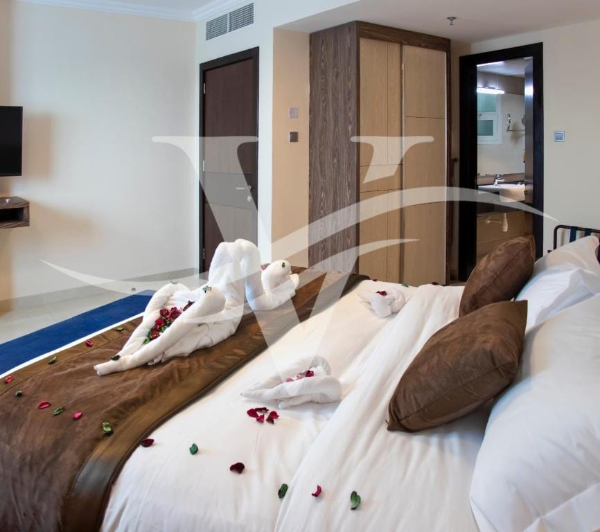 A well-appointed bedroom displays a spacious bed adorned with decorative pillows and arranged towels shaped like swans. Petals scatter across the bedding, contributing to a refined aesthetic. A wardrobe and a wall-mounted TV are visible in the background, while an ensuite bathroom door can be seen partially open.
