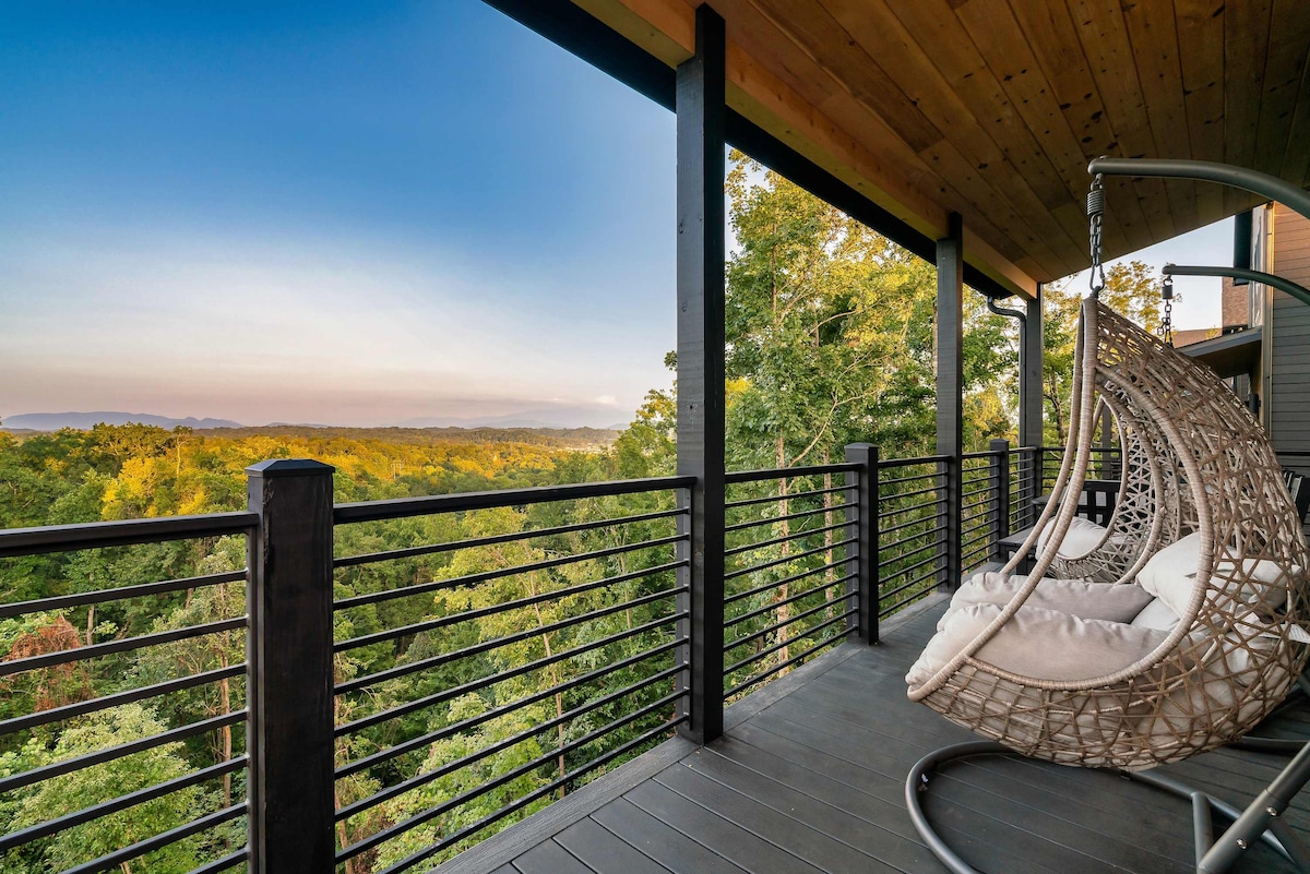 A spacious balcony offers expansive views of the surrounding landscape, showcasing lush greenery and distant mountains under a clear sky. A comfortable hanging chair is suspended, inviting relaxation while taking in the serene outdoor atmosphere.