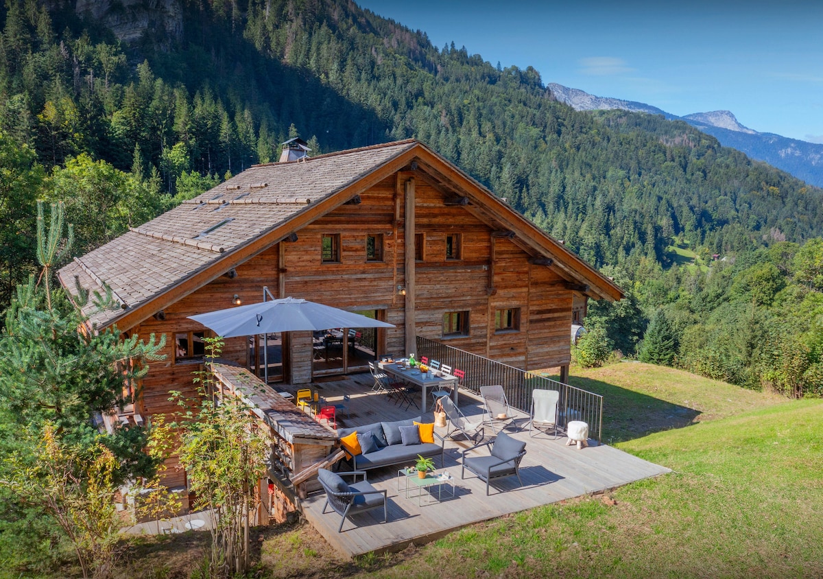 The chalet is nestled in a natural setting, featuring a spacious wooden terrace furnished with seating, including lounge chairs and a table. Umbrellas provide shade, while surrounding greenery and mountains enhance the charming atmosphere of the outdoor area.