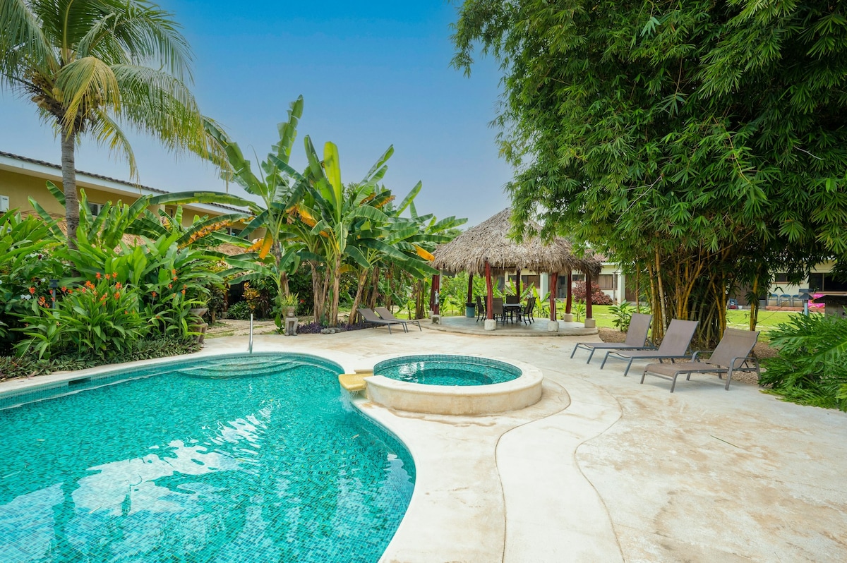 An inviting outdoor area is highlighted by a refreshing swimming pool surrounded by lush tropical plants. A thatched-roof shelter offers shade, while several lounge chairs are positioned nearby. A circular hot tub is visible next to the pool, enhancing the serene ambiance.