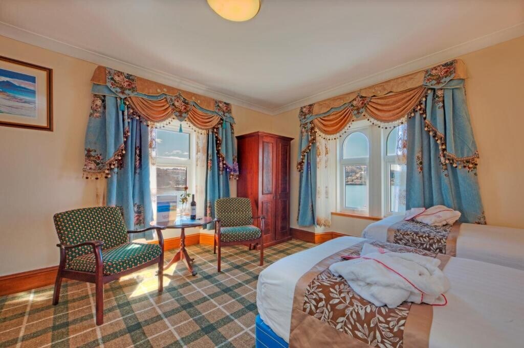 A Standard Twin Room is displayed with two twin beds, adorned with linens and towels. A coffee table and two chairs are arranged near a cabinet. Natural light filters through two windows dressed with patterned curtains, illuminating the warm interior.