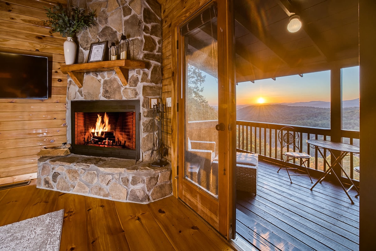 The interior of a cabin features a stone fireplace, with a cozy fire illuminating the room. A door leads to a balcony, where stunning mountain views can be seen at sunset. The space includes wooden accents and comfortable seating, enhancing the inviting atmosphere.