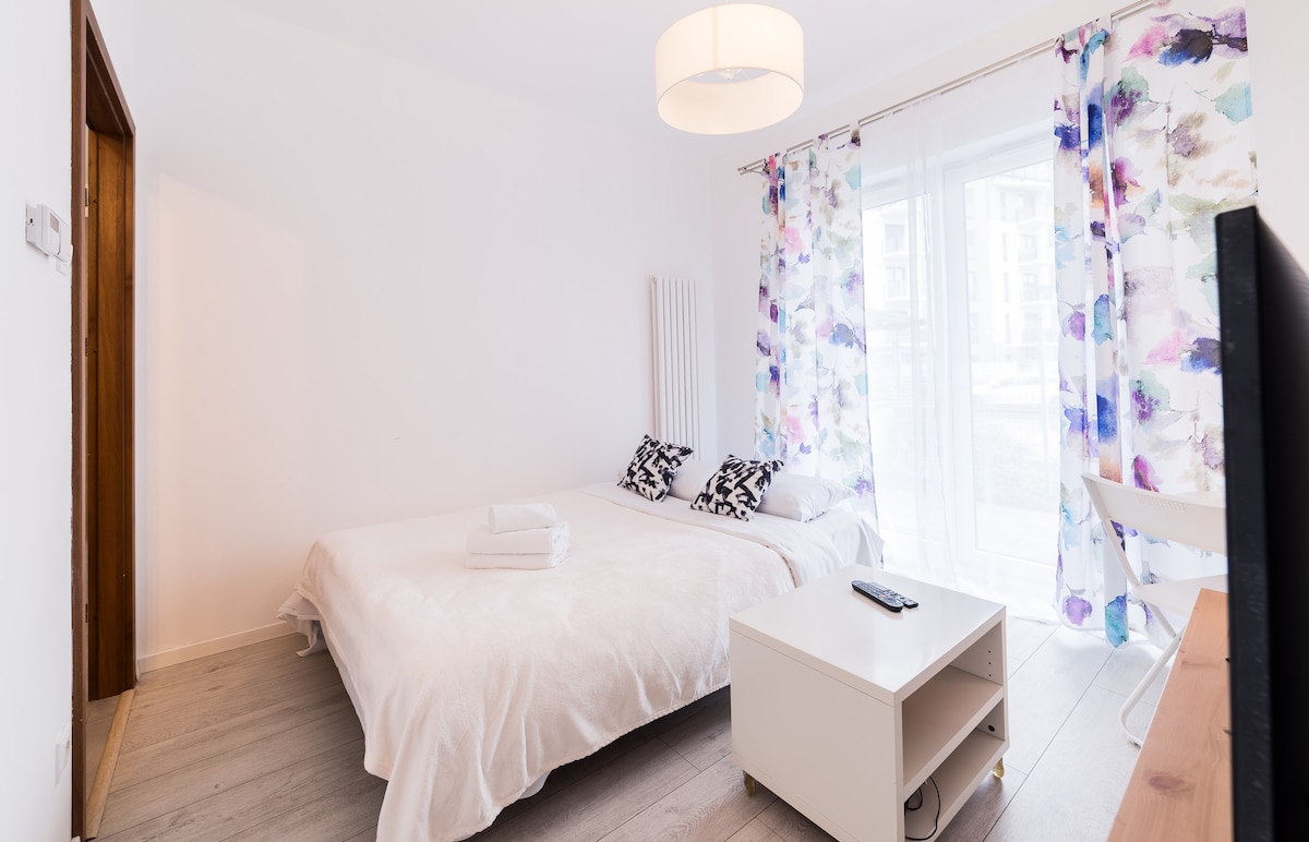 A light-filled bedroom features a spacious bed covered with a white blanket. Two decorative pillows are arranged at the head of the bed. Sheer, patterned curtains frame the large window, and a small white table with a remote control is positioned nearby.
