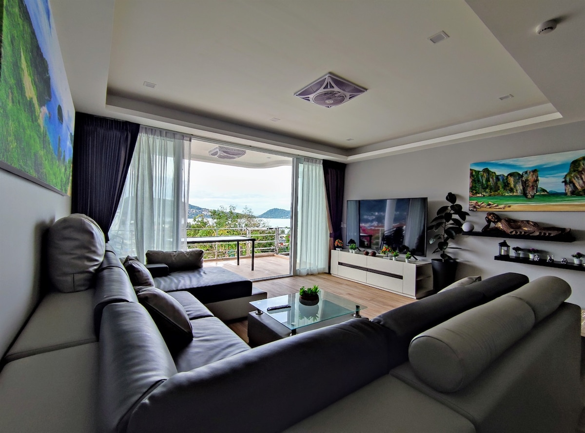 A modern lounge area is presented with a large sectional sofa facing a flat-screen television. Floor-to-ceiling windows allow natural light to fill the space, offering views of the outside greenery and sea. A glass table sits in the center, accompanied by decorative items.