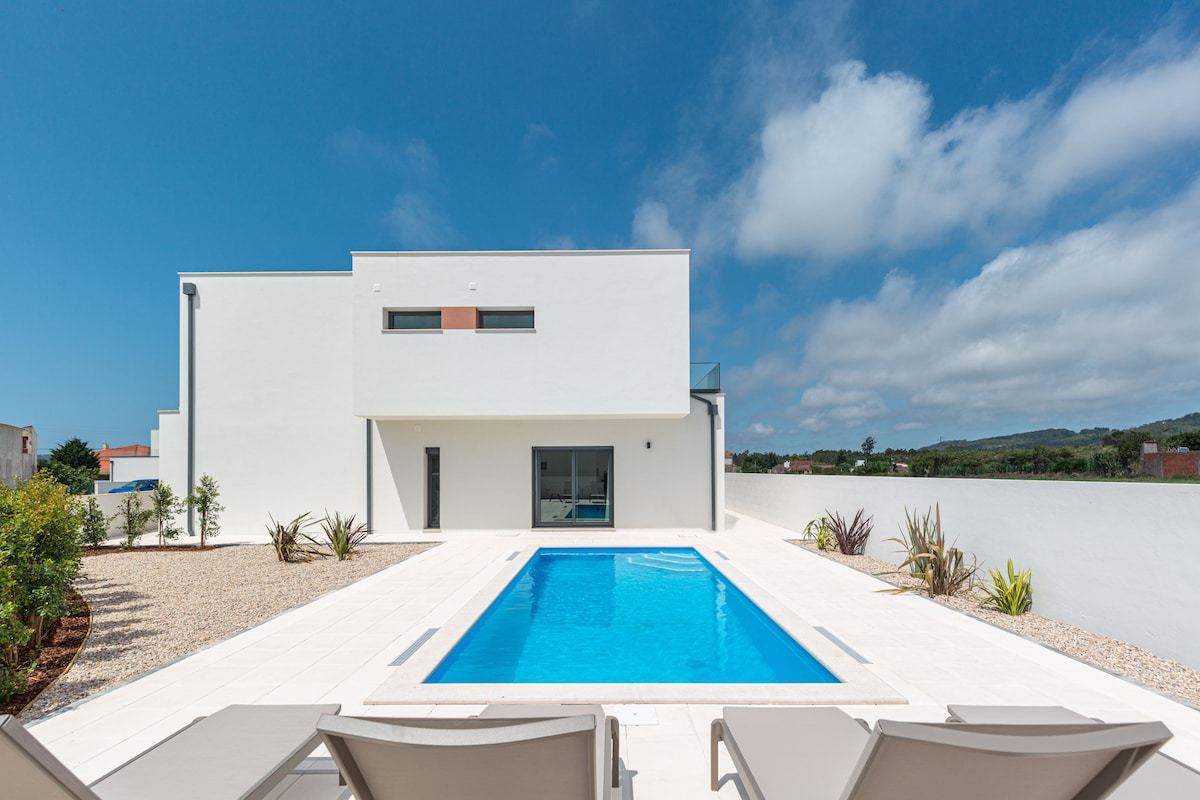 A contemporary villa is depicted with a clear blue swimming pool at the center. Sun loungers are positioned nearby, surrounded by landscaped areas featuring decorative plants. The exterior walls are smooth and white, set against a bright blue sky with scattered clouds.