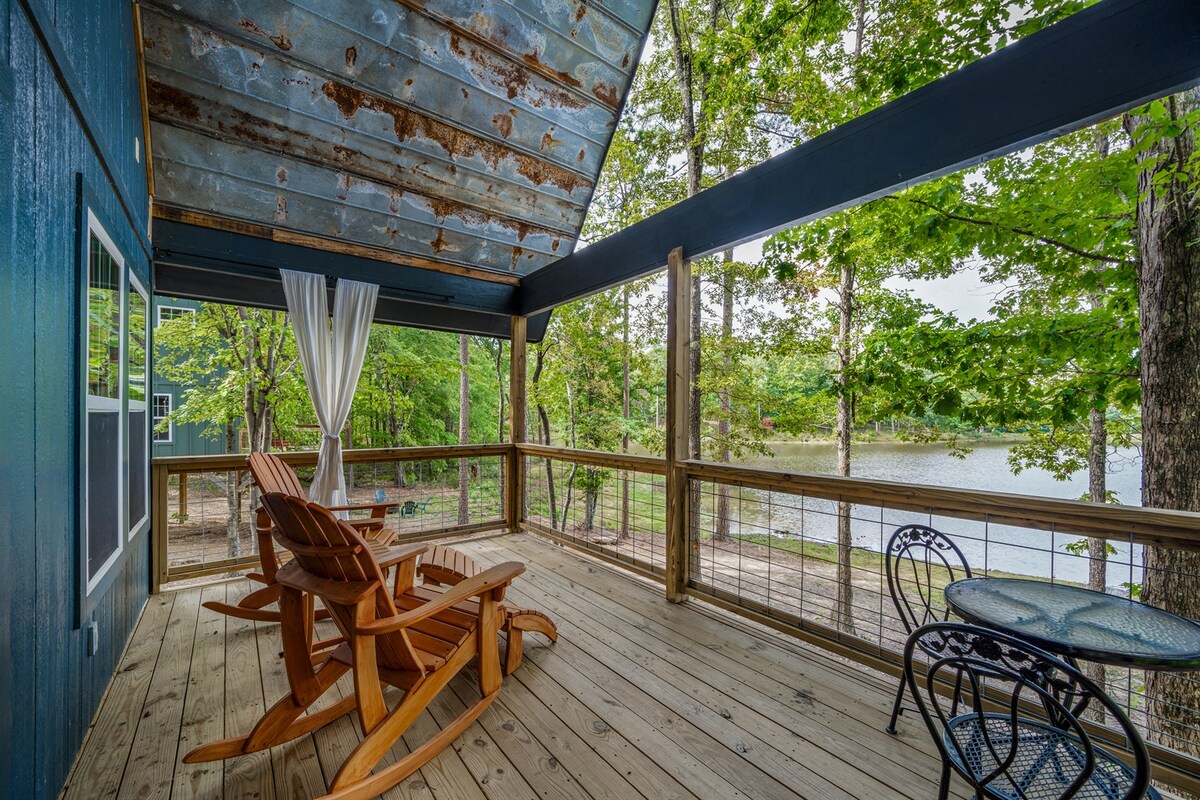An expansive deck overlooks Hidden Lake, furnished with two adirondack chairs and a small round table. Large windows allow natural light to illuminate the area, while trees provide a serene backdrop. A curtain softly billows in the breeze, adding a touch of tranquility to the setting.