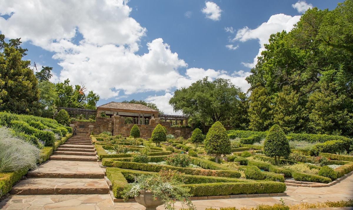 A well-maintained garden features neatly trimmed hedges and various greenery arranged in geometric patterns. Stone steps lead up to a raised area with a covered pavilion, surrounded by tall trees and expansive blue skies with scattered clouds.